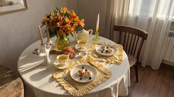 Mesa redonda con mantel, dos platos con postres, tazas de té, tetera, jarrón de flores, uvas, dos candelabros y una silla de madera. Luz del sol.