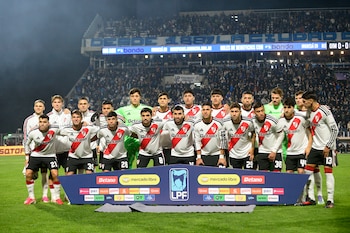 Dieciocho futbolistas con camisetas blancas y rojas posan en un campo de juego. Detrás hay un cartel de sponsors y tribunas con público