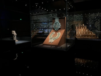 Una máscara de jade maya verde y un collar se exhiben en una vitrina iluminada en una sala de museo oscura, junto a figuras prehispánicas y una escultura de piedra