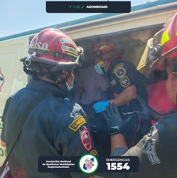Tres bomberos con uniformes, cascos rojos y guantes de protección asisten a una persona con mascarilla dentro de un vehículo de emergencia, en un entorno diurno