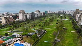 Vista aérea de campos de golf verdes, árboles, edificios urbanos altos, piscinas y canchas deportivas, con el mar en el horizonte de Lima bajo cielo nublado