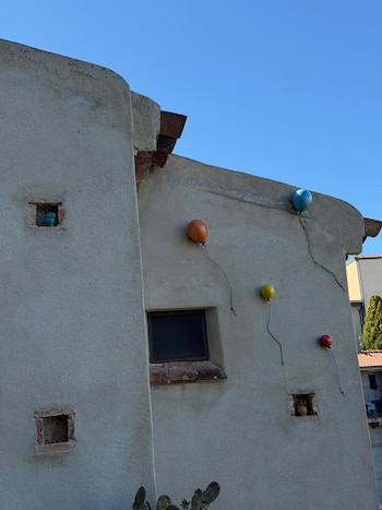 Fachada antiga colorida do edifício com quatro objetos em forma de balão: azul, laranja, amarelo e vermelho, fixados na parede sob um céu azul claro.