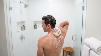 Vista posterior de un hombre que lava su espalda con una esponja en una ducha con azulejos blancos y mampara de vidrio.