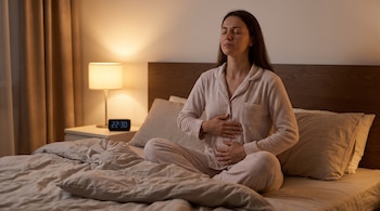 Mujer en pijama sentada en la cama con los ojos cerrados y las manos en el abdomen, junto a una mesa de noche con lámpara y reloj digital.