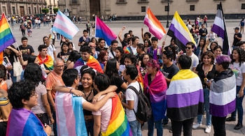 Grupo de personas diversas con banderas LGBTI+ (arcoíris, trans, bisexual, lésbica, no binaria, asexual) al aire libre en entorno urbano