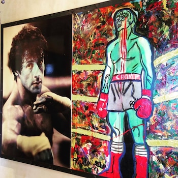 Fotografía en blanco y negro de un hombre con guantes de boxeo junto a un cuadro de un boxeador verde con guantes rojos y sangre goteando