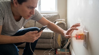 Un hombre mira un cargador de celular blanco conectado a un enchufe de pared. El cargador emite humo, luz roja y muestra signos de quemadura.