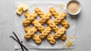 Vista superior de galletas trenzadas sobre papel blanco con mantequilla, semillas de sésamo, vainas de vainilla y cáscara de limón.