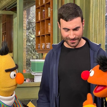 Un hombre de pelo oscuro y barba mira hacia abajo, con los Muppets amarillos Bert y el naranja Ernie. Al fondo hay estanterías y una ventana