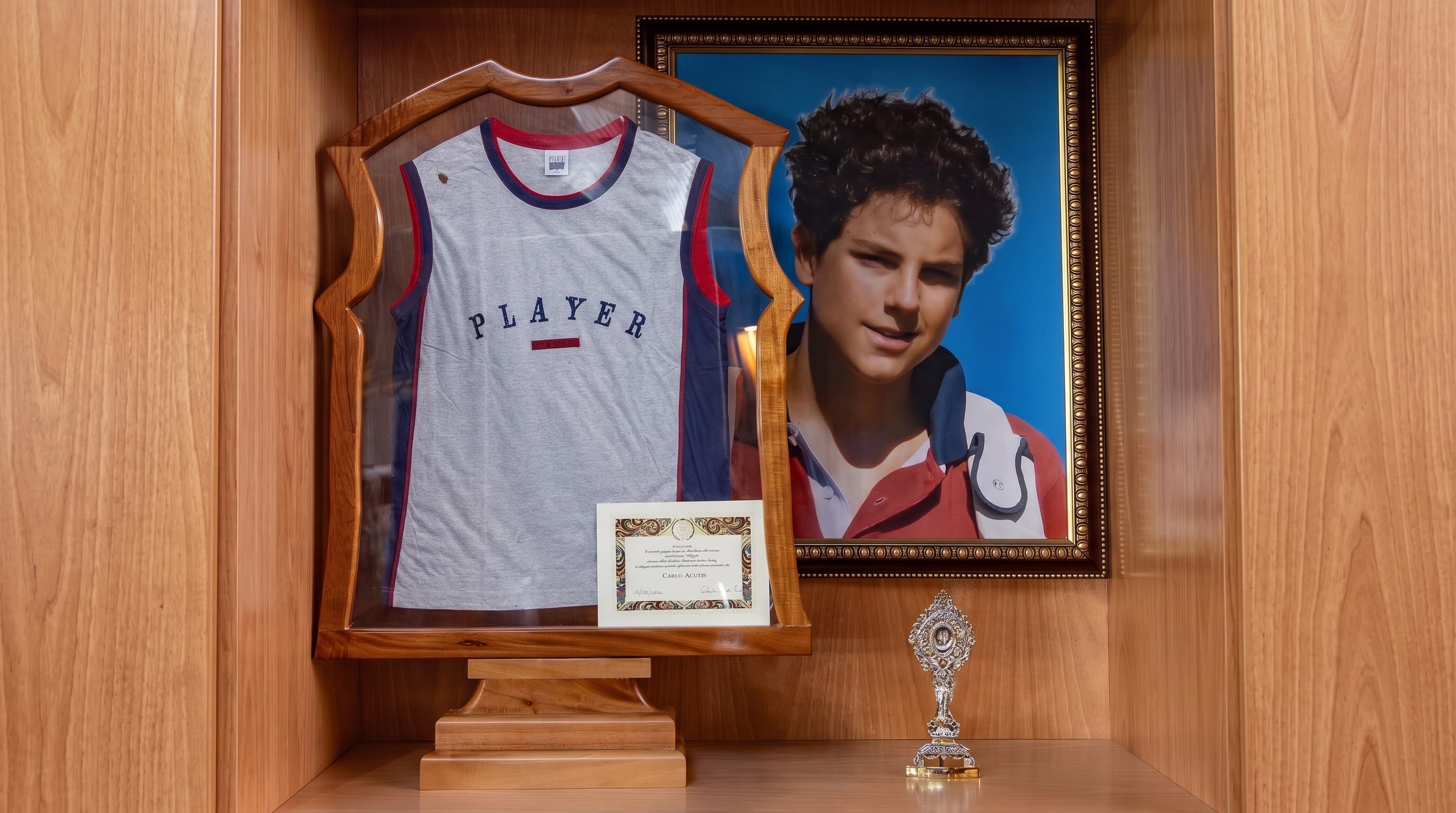 Un espacio memorial exhibe un jersey deportivo, un certificado de autenticidad y un retrato del beato Carlo Acutis junto a un relicario. (Imagen Ilustrativa Infobae)