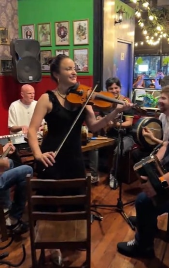 Mujer sonriente con violín toca en el centro, rodeada de otros músicos con instrumentos en un local con paredes rojas y verdes, y luces cálidas