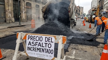 Un camión volquete descarga asfalto caliente en una calle, mientras dos obreros lo esparcen. Un cartel blanco anuncia el incremento del precio del asfalto.