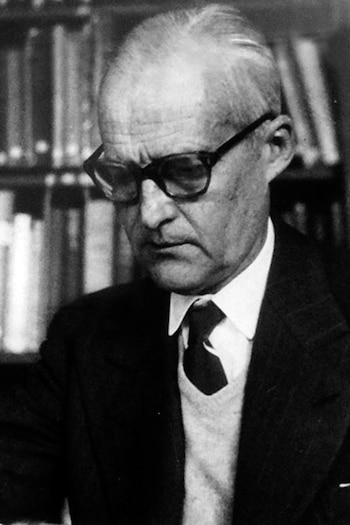 Fotografía en blanco y negro de un hombre con anteojos, camisa, corbata, suéter y saco, ubicado delante de una biblioteca con libros