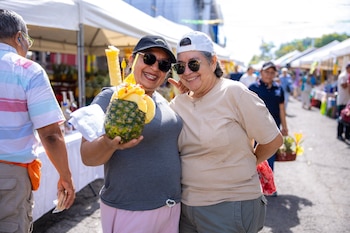 Hombre y mujer asiáticos sonriendo en primer plano, la mujer sostiene un letrero con forma de piña que dice "Feria de la Piña 2026 Santa María Ostuma"