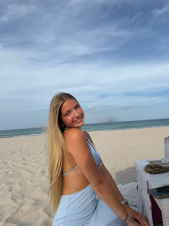 Joven con cabello rubio largo sonríe sentada en la arena de una playa con el mar y el cielo de fondo