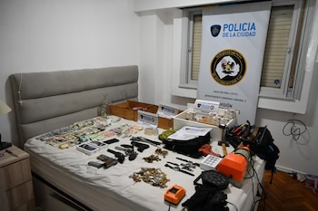 Una cama con dinero en efectivo, armas de fuego, municiones, teléfonos celulares, un rosario y herramientas, con un cartel de la Policía de la Ciudad al fondo