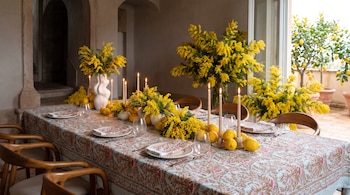 Mesa con mantel estampado, vajilla, copas, velas encendidas, limones y arreglos de mimosas amarillas, junto a limoneros en macetas.