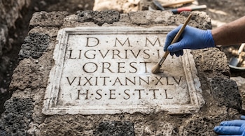 Manos con guantes azules usan un pincel para limpiar las letras grabadas en una placa de piedra antigua rodeada de bloques oscuros.