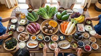 Vista aérea de una mesa de madera con personas compartiendo una comida de la dieta Okinawa: frutas, vegetales, pescado, tofu, arroz, frutos secos y bebidas.