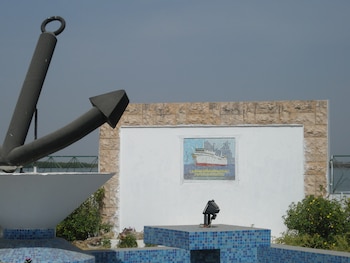 Una gran ancla negra junto a una pared blanca con un mosaico que representa a un barco de pasajeros sobre azulejos azules