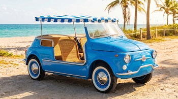 Coche vintage azul estilo Fiat Jolly con capota a rayas y asientos de mimbre estacionado en una playa de arena clara, con el mar azul y palmeras.