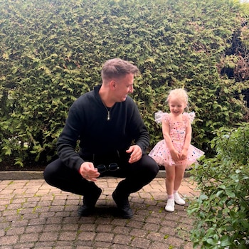 Maxi Lopez, vestido de negro, agachado frente a una niña rubia con vestido de ballet rosa con puntos de colores, ambos sonriendo frente a un seto verde