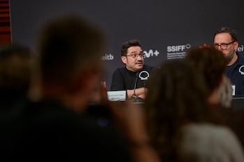 Juan Antonio Bayona habla ante un micrófono tras una mesa con su cartel de identificación y un panel con logotipos detrás