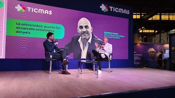 Dos hombres sentados en un escenario de madera conversan frente a una gran pantalla LED púrpura que muestra el logo de TICMAS, un retrato y texto sobre desarrollo estratégico