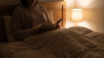 Una persona en la cama lee un libro con una lámpara de mesa encendida a la derecha, debajo de una colcha de color claro y con almohadas.