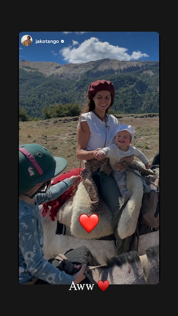 Una mujer con boina roja y un bebé van montados a caballo junto a un niño con casco en un paisaje de montaña