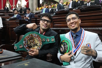 Dos boxeadores sonrientes sostienen cinturones de campeonato WBC de color verde y dorado, sentados en un recinto oficial con personas en segundo plano