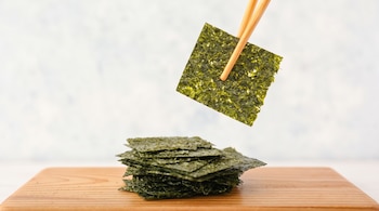 Unos palillos de madera sostienen una lámina cuadrada de alga nori sobre una pila de algas en una tabla de madera.