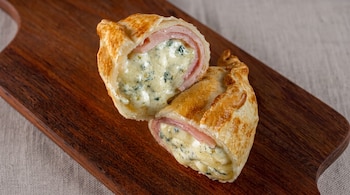 Una empanada cortada en dos partes con relleno visible de queso y jamón sobre una tabla de madera y un mantel gris.