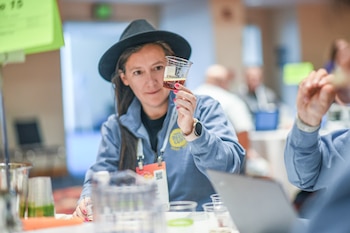 Mujer con sombrero y sudadera azul sujeta un vaso de cerveza, con esmalte de uñas rosa y reloj. Hay otros vasos y personas borrosas de fondo