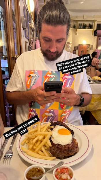 Hombre barbudo con pelo recogido y camiseta de los Rolling Stones mirando su móvil, con plato de carne, patatas fritas y huevo frito en primer plano