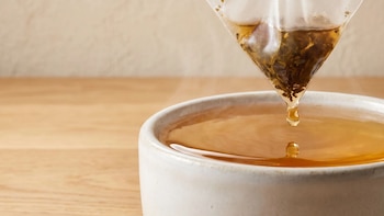 Una bolsita de té piramidal gotea sobre una taza blanca con infusión caliente encima de una mesa de madera