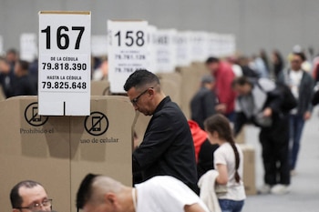 Un hombre con gafas y chaqueta oscura vota en una cabina de cartón en un centro electoral, con carteles que prohíben el uso de celulares