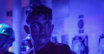 Primer plano de un joven con cabello oscuro, mirando a la derecha con expresión seria, iluminado por una luz azul; en el fondo, figuras borrosas y equipos