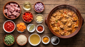 Ingredientes para guiso de cerdo con arroz: carne en cubos, verduras, arroz, especias y caldo. Una olla humeante del plato terminado sobre mesa de madera.