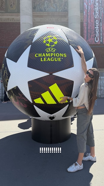 Mujer con gafas de sol sonríe abrazando un balón gigante blanco y negro de la UEFA Champions League, en un espacio abierto con un edificio de arquitectura clásica al fondo