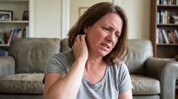 Mujer con camiseta gris se rasca el cuello cerca de la oreja con la mano derecha. Su rostro tiene ceño fruncido. Sofá y estanterías se ven borrosos detrás.