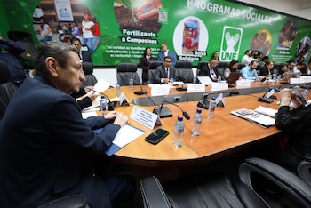 Personas sentadas alrededor de una mesa con micrófonos y letreros mientras un grupo de periodistas toma fotografías