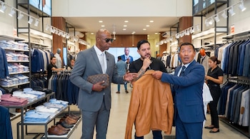 En una tienda de ropa, tres hombres, incluyendo uno con traje gris y gafas, y otro con traje azul, sostienen y examinan una chaqueta de cuero marrón.