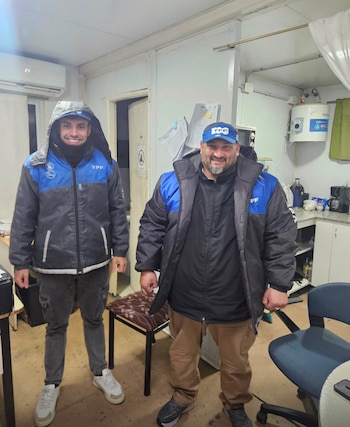 Dos hombres sonríen, uno con capucha, ambos con gorra y chaqueta de YPF, dentro de una habitación con aire acondicionado, calentador de agua y muebles
