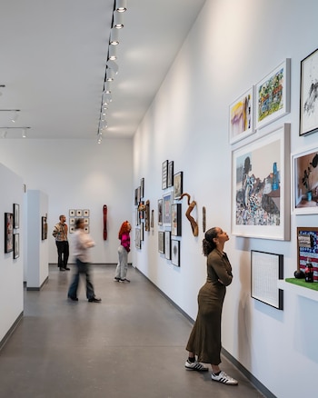 Un grupo de personas observa varias obras de arte en una galería moderna de paredes blancas y suelo gris. Una mujer mira hacia arriba a un cuadro grande
