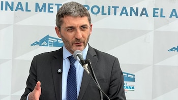 Hombre con gafas, barba y traje oscuro habla en un micrófono en un evento. Detrás, una pared blanca con texto azul parcial y gráficos de edificios