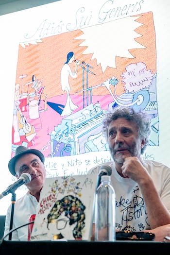Dos hombres sentados en una mesa con micrófonos; el de la derecha es Fito Páez, y detrás de ellos se ve una gran pantalla con una ilustración de la banda Sui Generis