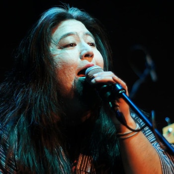 Primer plano de una persona de pelo largo oscuro con canas, cantando apasionadamente en un micrófono plateado, vestida con una camisa a rayas