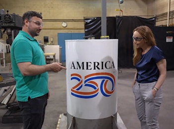 Un hombre y una mujer con gafas de seguridad miran una cápsula del tiempo cilíndrica blanca con el logo "AMERICA 250" en un taller