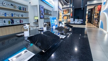 Interior de una tienda de electrónica. Mostrador negro con tabletas y dispositivos en exhibición. Estantes con accesorios. Un pasillo de centro comercial se ve al fondo